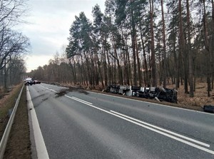 Zdjęcie kolorowe: zdjęcie jezdni. Z prawej strony w rowie widoczna wywrócona cysterna, na środku drogi czerwony samochód.