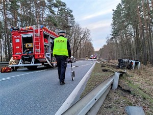 Zdjęcie kolorowe: umundurowany policjant ruchu drogowego podczas wykonywania czynności na miejscu wypadku drogowego. W tle widoczny wóz strażacki, miejsce zdarzenia.