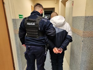 Zdjęcie kolorowe: umundurowany policjant z zatrzymanym mężczyzną, który ma założone kajdanki na ręce, które trzyma z tyłu