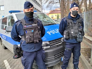 Zdjęcie kolorowe: dwóch umundurowanych policjantów, w tle widoczny radiowóz policyjny.