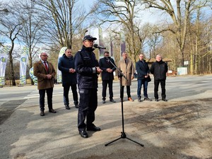 Zdjęcie kolorowe: umundurowany policjant przemawiający przy mikrofonie, w tle widoczni mężczyźni