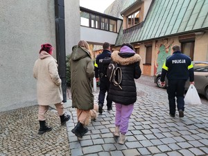 Zdjęcie kolorowe: na pierwszym planie widoczne trzy kobiety. Dalej dwóch umundurowanych policjantów którzy niosą torby, plecaki.