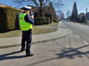 Zdjęcie kolorowe: umundurowany policjant ruchu drogowego podczas pomiaru prędkości przy drodze