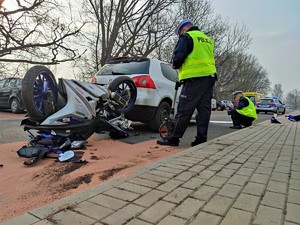 Zdjęcie kolorowe: dwóch umundurowanych policjantów ruchu drogowego podczas czynności na miejscu wypadku. Widoczny wywrócony motocykl. W tle srebrny samochód osobowy.