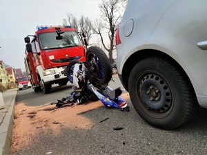 Zdjęcie kolorowe: widoczny wywrócony motocykl, w tle wóz straży pożarnej