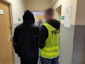 Zdjęcie kolorowe: policjant w kamizelce odblaskowej z napisem &quot;Policja&quot; wraz z zatrzymanym mężczyzną w korytarzu