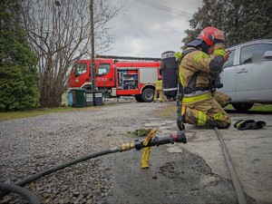 Zdjęcie kolorowe: na pierwszym planie wąż strażacki, dalej widoczny strażak który klęcząc zdejmuje maskę. W tle wóz straży pożarnej