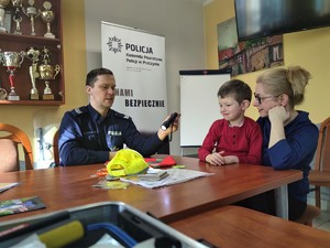 Zdjęcie kolorowe: umundurowany policjant siedzi przy stole, obok chłopiec z mamą. Policjant świeci latarką na odblaskową czapkę z daszkiem. Na stole widoczne różne książeczki i kolorowanki.