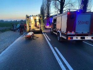 Zdjęcie kolorowe: z prawej strony widoczny wóz straży pożarnej, z lewej otwartą karetkę pogotowia, przed nią nosze.