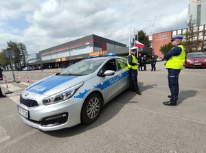 Zdjęcie kolorowe: oznakowany policyjny radiowóz z włączonymi sygnałami świetlnymi, dwóch policjantów ruchu drogowego przed kopalnią KWK Pniówek