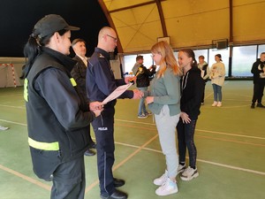 Zdjęcie kolorowe: policjant gratuluje dziewczynce. Obok widoczni strażacy.