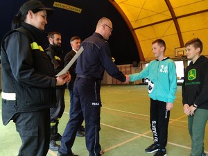 Zdjęcie kolorowe: policjant gratuluje uczestnikowi. Obok widoczni strażacy.