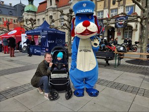 Zdjęcie kolorowe: policyjna maskotka wraz z kobietą i dzieckiem w wózku