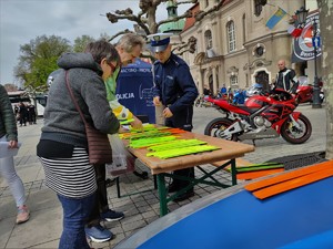 Zdjęcie kolorowe: umundurowany policjant przy stoliku z odblaskami, obok przechodzą ludzie