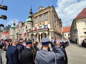 Zdjęcie kolorowe: komendant policji i straży pożarnej salutują podczas gdy reszta uczestników stoi na baczność, w tle widoczny budynek urzędu miasta w Pszczynie.