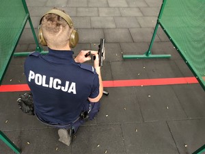 Zdjęcie kolorowe: umundurowany policjant klęczy na jednym kolanie i prezentuje rozładowaną broń