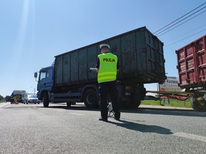 Zdjęcie kolorowe: umundurowany policjant ruchu drogowego podczas czynności na miejscu zdarzenia, w tle widoczny pojazd ciężarowy