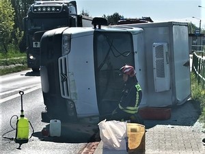 Zdjęcie kolorowe: samochód typu bus przewrócony na bok, przed nim klęczy strażak