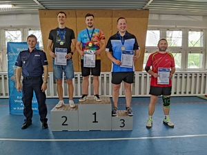 Zdjęcie kolorowe: na podium stoi trzech mężczyzn z dyplomami i medalami, z lewej strony stoi umundurowany policjant, z prawej mężczyzna z dyplomem.