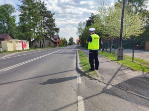 Zdjęcie kolorowe: umundurowany policjant ruchu drogowego podczas pomiaru prędkości przy drodze.