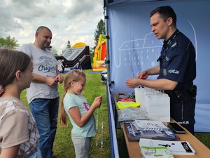 Zdjęcie kolorowe: na pierwszym planie dziewczynka i umundurowany policjant który daje jej odblask, dalej widoczne osoby