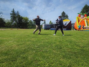 Zdjęcie kolorowe: dwóch mężczyzn naprzeciwko siebie, osoby podczas pokazu sztuk walki, jeden z mężczyzn bierze zamach pałką, drugi się broni