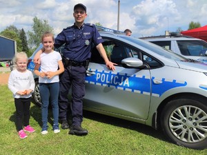 Zdjęcie kolorowe: dwie dziewczynki wraz z umundurowanym policjantem pozują do zdjęcia, w tle radiowóz