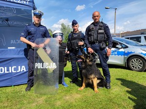 Zdjęcie kolorowe: zdjęcie pozowane. Widoczny policjant ruchu drogowego, policjantka prewencji i przewodnik psa służbowego wraz z czworonogiem, na środku stoi dziewczynka w kamizelce i kasku policyjnym