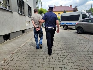 Zdjęcie kolorowe: umundurowany policjant prowadzi zatrzymanego mężczyznę