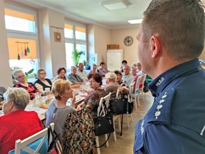 Zdjęcie kolorowe: na pierwszym planie pagony z munduru policjantka, w tle widoczni seniorzy przy stole