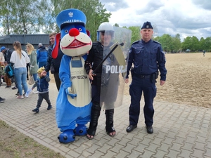 Zdjęcie kolorowe: policyjna maskotka sznupek, nastolatek w stroju ochronnym z tarczą, pałką i kasku oraz umundurowany policjant podczas pozowanego zdjęcia