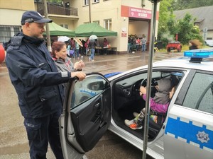 Zdjęcie kolorowe: umundurowany policjant stoi obok radiowozu w którym siedzi dwoje dzieci, obok widoczna kobieta która robi im zdjęcie