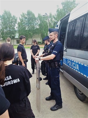 Zdjęcie kolorowe: umundurowani policjanci na tle radiowozu stoją naprzeciwko uczniów klasy mundurowej i mówią