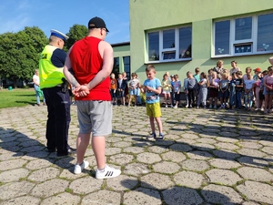 Zdjęcie kolorowe: umundurowany policjant ruchu drogowego stoi naprzeciw grupy dzieci. Z przodu widoczny chłopczyk