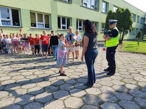 Zdjęcie kolorowe: dziewczynka podaje kobiecie laurkę w kształcie kwiatka, obok stoi umundurowany policjant. W tle widoczne dzieci wraz z opiekunami