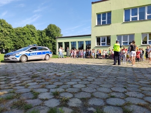 Zdjęcie kolorowe: umundurowany policjant stoi naprzeciwko grupy dzieci z opiekunami, obok niego kobieta.