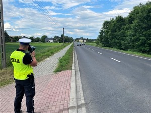 Zdjęcie kolorowe: umundurowany policjant podczas pomiaru prędkości przy drodze