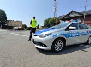 Zdjęcie kolorowe: widoczny umundurowany policjant, który zabezpiecza trasę przejazdu kolarzy, z prawej strony oznakowany radiowóz policyjny.