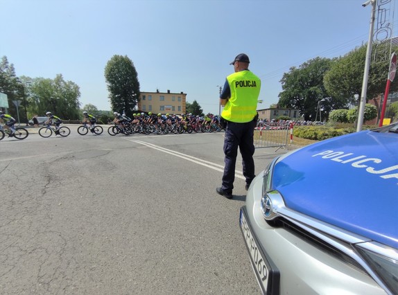 Zdjęcie kolorowe: umundurowany policjant podczas zabezpieczenia przejazdu kolarzy. W tle widoczni rowerzyści.
