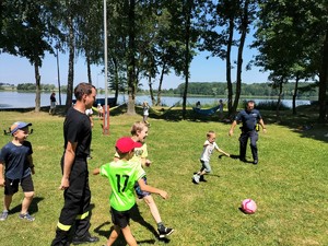 Zdjęcie kolorowe: policjanci i strażacy na boisku grają z dziećmi w piłkę nożną.