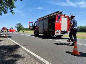 Zdjęcie kolorowe: miejsce zdarzenia drogowego, widoczne rozbite pojazdy, policjant, strażacy.