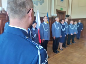 Zdjęcie kolorowe: na pierwszym planie widoczne pagony umundurowanego policjanta, w tle grupa policjantów stoi w szeregu.