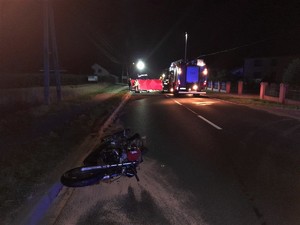 Zdjęcie kolorowe: miejsce zdarzenia drogowego, widoczny uszkodzony motocykl, w tle wozy straży pożarnej.