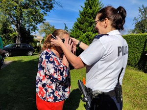 Zdjęcie kolorowe: umundurowana policjantka pomaga starszej pani założyć google.