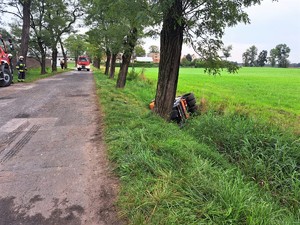 Zdjęcie kolorowe: miejsce zdarzenia drogowego. Widoczne wozy straży pożarnej, a także wywrócony walec drogowy w przydrożnym rowie.