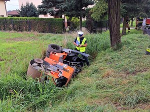 Zdjęcie kolorowe: wywrócony walec drogowy leży w przydrożnym rowie, obok stoi umundurowany policjant ruchu drogowego.