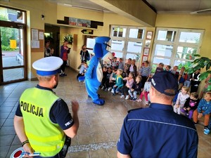 Zdjęcie kolorowe przedstawiające dwóch umundurowanych policjantów, policyjną maskotkę i grupę dzieci w szkole.