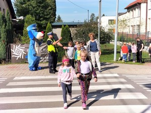 Zdjęcie kolorowe przedstawiające dzieci przechodzące przez przejście dla pieszych w obecności umundurowanego policjanta ruchu drogowego.