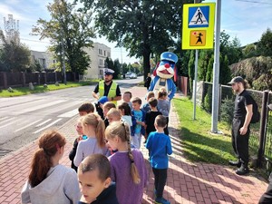 Zdjęcie kolorowe przedstawia grupę dzieci wraz z policjantem i policyjna maskotką przed oznakowanym przejściem dla pieszych.