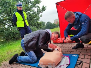 Zdjęcie kolorowe przedstawiające mężczyznę podczas ćwiczeń pierwszej pomocy. Widoczny także strażak i policjant.